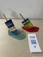 Photo showing both resin melting popsicle sculptures with popsicle sticks angled, placed on a light surface, with a MaxSold bidding number.