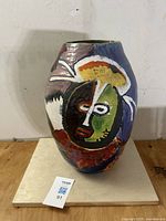 Front view of the pottery vase showing one side with an abstract face in black, green, white, yellow, and red on a purple background.