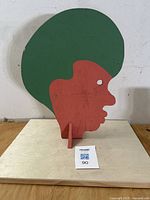 Front view of painted folk art wooden sculpture showing profile with large green hair and red face, standing on a wooden base.