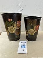 Front view of two antique tole flower pails showing hand-painted floral motifs on the dark metal surface with a MaxSold tag in front.