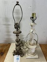 Two vintage accent lamps side by side on a wooden surface against a plain indoor wall; the left lamp is distressed gilt with ornate scrollwork base, the right lamp is white marble with natural veining and baluster shape.