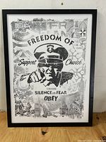 Framed black and white screen print showing the entire artwork with figure and textual elements, placed on a wooden surface.