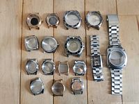 Top view of all 16 watch cases arranged on wooden surface showing variety of shapes, colors, and attached bracelets on two cases.