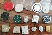 Top-down view of all watch movements and dials arranged on wood surface showing various shapes, sizes, colors, and brands.
