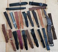 Overhead view of 23 vintage watch straps arranged on a wooden surface. Straps include various colors such as black, brown, tan, burgundy, and grey with assorted textures and buckle styles.