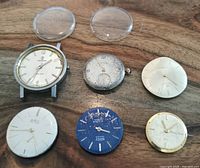 Overview of six men's watch movements and watch faces on wooden surface with two clear watch crystals