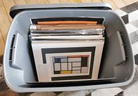 Plastic crate filled with art prints, mostly individually wrapped in clear protective sleeves.