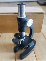 Front angle view of the black Birks microscope with metal body, showing its U-shaped stand with stage platform and wooden box in background.