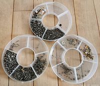 Three plastic compartment organizers with various small metal watch parts including screws, stems, crowns, and small pins.