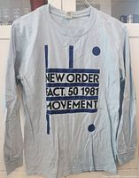 Light blue long sleeve t-shirt with navy blue graphic text in front showing 'NEW ORDER FACT. 50 1981 MOVEMENT'. Shirt has ribbed cuffs, slightly wrinkled but in excellent condition.