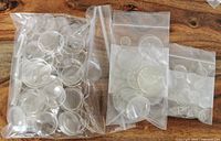 Three resealable plastic bags containing various sizes of clear round acrylic watch crystals laid on a wooden surface.
