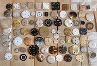 Wide overhead shot showing entire assortment of mostly round and some square watch dials, laid out on a wooden surface.