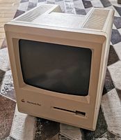 Front view of the vintage Macintosh Plus computer showing the CRT screen and floppy disk drive with Apple logo.