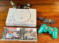 PlayStation 1 console with controller, two game cases, and cables laid out on wooden surface