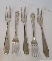Five antique silver forks displayed side by side showing the front side with decorative floral patterns on handles.