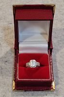 The ring displayed inside a red velvet and satin jewelry box, showing the full ring design and setting.