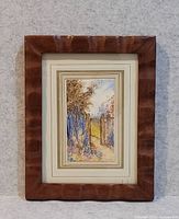 Frontal image of the framed antique watercolour painting showing a rustic outdoor scene with a pathway, trees, and fences in soft muted colors.