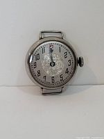 Front view of an antique wristwatch showing the dial with black Arabic numerals and a floral engraved center.