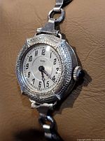 Front close-up of antique Relo wristwatch showing silver engraved case, black Arabic numerals on a silver dial, and black cabochon crown on right.
