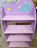 Front view showing 4-tier lavender purple kids bookshelf with teal unicorn and yellow stars painted on top back panel.