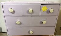 Front view of six drawer storage box showing three small drawers on top, two medium drawers in the middle and one large drawer at bottom, each with silver knobs. Yellow flower sticker on right top drawer.