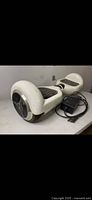 Side angle view of white hoverboard with black foot pads and one wheel. Black charging adapter plugged in with cord shown.