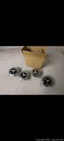 Four Mercedes Benz wheel caps displayed unmounted with original box in background, each wrapped in protective plastic.