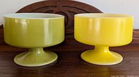 Two milk glass footed bowls in yellow and green, shown side by side on a wooden surface.