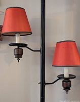 View of the tension pole lamp with two orange lampshades attached on black metal arms extending from the vertical pole. Wooden and metal accents at lamp holders visible.
