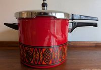 Side view of the red enamel Sicomatic pressure cooker showing decorative orange and black geometric leaf pattern at base, with black plastic handles and stainless steel lid topped with pressure release valve.