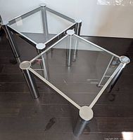 Two square side tables with chrome legs and glass tops on dark hardwood floor, visible light scratches on glass surfaces