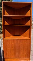 Full view of the teak corner cabinet showing three open shelves on top and closed double-door cabinet below with teak wood grain finish.