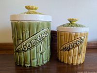 Photo showing two vintage ceramic canisters labeled 'Cookies' (green) and 'Coffee' (tan) with bamboo texture and leaf design lids with flower knobs.