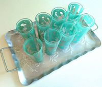 Set of eight turquoise tall glasses with white musical-themed etched designs arranged on a metal serving tray with floral embossing and handles.