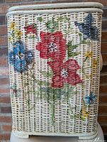 Close-up photo of vintage wicker laundry basket showing painted floral design with red, blue, yellow flowers on natural wicker