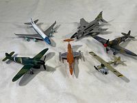 Top view of seven diecast toy planes, showing variety of WWII and modern styles including commercial jet and fighter planes on white fabric.