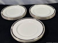 Seven white Aynsley bone china plates with black and gold edge decoration stacked on a black surface.
