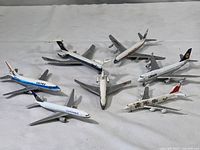 Seven vintage metal toy airplanes arranged on a white surface, showing different airline liveries including Lufthansa and JAL. Some planes lack landing gear.