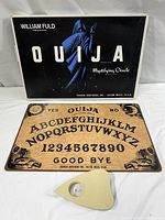 Photo showing the Ouija board laying flat with the original box behind and plastic planchette in front.