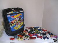 Plastic black Hot Wheels carrying case with racing car graphics and text 'Challenge Accepted' that holds 100 cars, positioned on a white shelf next to multiple Hot Wheels cars spread out.