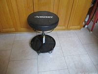 Side view of the black Husky tool stool showing the padded seat, adjustable height mechanism, lower tray shelf, and caster wheels.