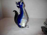 Full side view of Murano glass cat figurine showing blue and clear glass body with black tail and small label on the front foot.