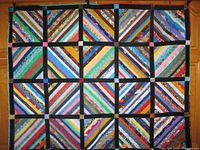 Full view of handmade patchwork quilt hanging, displaying colorful diagonal fabric strips in square blocks separated by black sashing with accent squares.