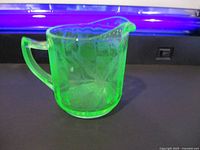 Front side view of small green uranium glass jug under UV light, showing handle and pouring spout.