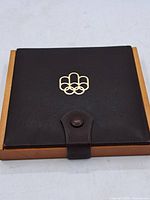 Closed brown official case with Olympic rings emblem on front and snap button closure.
