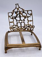 Front view of antique brass trivet showing the ornate folding stand and rectangular base with four ball feet.
