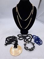 Display of six assorted necklaces including two pearl-style with clear and black beads and four beaded necklaces in black and blue colors, one with a large round mother-of-pearl pendant.
