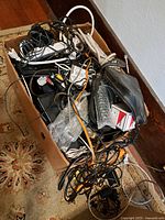 Box filled with assorted tangled electrical cables, wires, adapters, and a black pouch