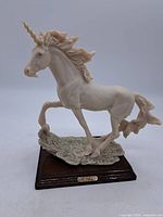 Front view of white resin unicorn figurine with one front hoof raised on textured base and wooden stand