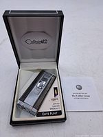 Colibri silver-tone Elite Flint lighter in open black cushioned box with warranty card beside it.